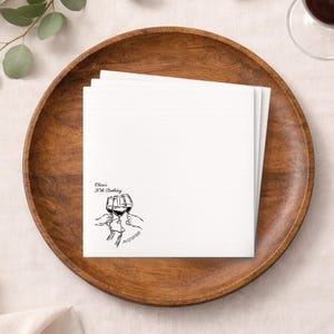 Puede incluir: Una pila de servilletas cuadradas blancas con un dibujo lineal negro de tres copas de vino brindando. El texto "Olivia's 30th Birthday" y la fecha 10/07/2020 también están impresos en las servilletas. Las servilletas están sobre una bandeja redonda de madera.