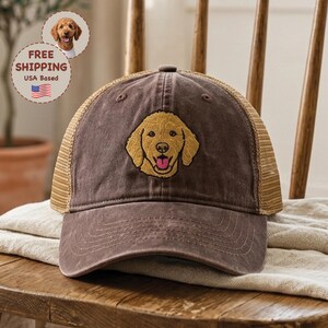 May include: A brown baseball cap with a mesh back and a golden retriever dog embroidered on the front. The dog has a pink tongue and is smiling. The hat is sitting on a wooden chair.