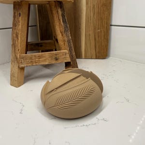 May include: A tan-colored, decorative object with a feather design, resting on a white countertop. A small wooden stool and a wooden cutting board are in the background. The object has a textured surface.