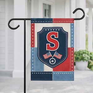 May include: A garden flag with a patriotic design, featuring a red "S" on a blue shield, two American flags, and stars. The flag has a patchwork pattern in red, white, and blue. It is displayed on a black metal garden stand.