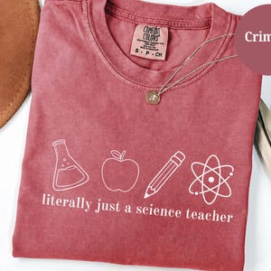 May include: A crimson short-sleeve t-shirt featuring a white graphic print of a laboratory flask, an apple, a pencil, and an atom symbol. Below the icons, the text reads literally just a science teacher in a white sans-serif font.
