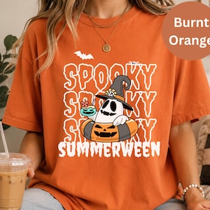 May include: A burnt orange t-shirt featuring a graphic of a ghost wearing a witch hat and sitting in a pumpkin-shaped pool float. The ghost holds a tropical drink. The text reads SPOOKY SPOOKY SPOOKY SUMMERWEEN in white, wavy, outlined letters.