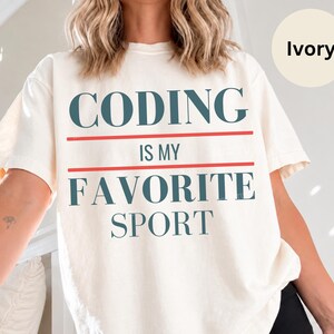 Op de afbeelding: Een ivoorkleurig T-shirt met korte mouwen en de donkerblauwe tekst "CODING IS MY FAVORITE SPORT" in een vetgedrukt lettertype. De tekst wordt gescheiden door twee horizontale rode lijnen en het shirt wordt gedragen met een zwarte short.