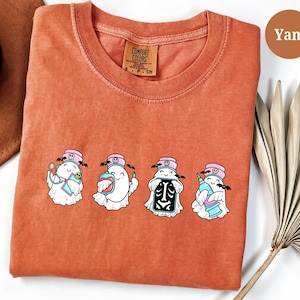 Pode incluir: Uma t-shirt de manga curta cor de ferrugem com um gráfico de quatro fantasmas brancos vestidos como pessoal médico. Cada fantasma usa uma touca de enfermeira rosa com um ícone de caveira e segura itens médicos como estetoscópio e seringa.