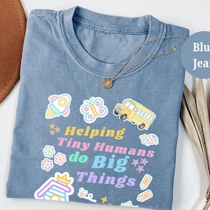 Puede incluir: Camiseta azul vaquero con la frase "Helping Tiny Humans do Big Things" y gráficos coloridos. Un collar dorado con un colgante descansa sobre la camiseta. Es una prenda de vestir informal.