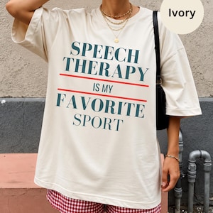 May include: An ivory oversized t-shirt featuring the dark teal text SPEECH THERAPY IS MY FAVORITE SPORT in a bold serif font, separated by two horizontal red lines. The shirt has short sleeves and a crew neckline, worn with red and white gingham shorts.