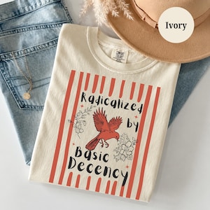 Könnte beinhalten: Ein elfenbeinfarbenes T-Shirt mit einer roten Vogelgrafik, floralen Akzenten und dem Text "Radicalized by Basic Decency" in schwarzer Schrift. Das Design ist von vertikalen roten Streifen umrahmt. Das T-Shirt hat einen Rundhalsausschnitt und ein Comfort Colors Etikett.