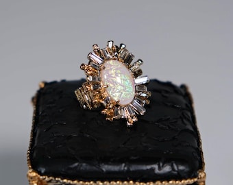 Puede incluir: Anillo dorado con una gran piedra central de ópalo ovalada, rodeada de cristales transparentes en forma de baguette y redondos. El anillo descansa sobre un pequeño cojín negro texturizado con ribetes dorados.