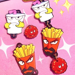 May include: A collection of cartoon character keychains featuring a white cup with a pink straw, a red fry container, and a red meatball. The keychains have black outlines and yellow accents. The background is pink with white starbursts.