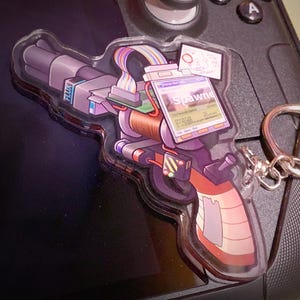 Puede incluir: Un llavero acrílico transparente con forma de pistola de rayos futurista. La pistola presenta detalles morados, naranjas y grises, con una pantalla que dice "Spawn". La palabra "FLUX" es visible. Se adjunta un llavero plateado.