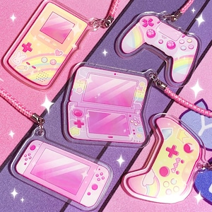 Peut inclure: Collection de breloques de consoles de jeux vidéo roses et jaunes, dont une Game Boy Color, une Nintendo 3DS, une Nintendo Switch et deux manettes. Chaque breloque a un cordon rose.