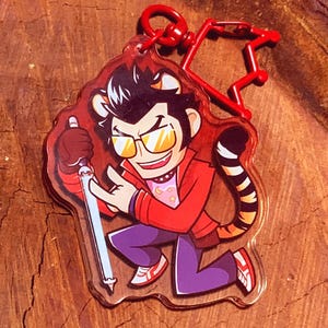 May include: A clear acrylic keychain featuring a cartoon character with tiger-striped ears and tail, wearing a red jacket, purple pants, and sunglasses. The keychain has a red clasp and a red star-shaped connector.