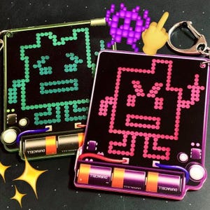 May include: Two retro-style keychains with pixelated character designs. One keychain features green lights, the other pink. Both have a black background, silver key rings, and Duracell batteries. A purple pixelated alien and a yellow hand emoji are also present.