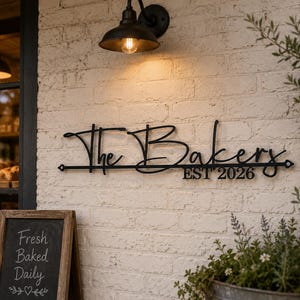 Puede incluir: Un letrero de pared de metal negro que presenta el texto en cursiva The Bakers y el texto impreso EST 2026. El letrero está montado en una pared de ladrillo blanco, colocado horizontalmente con puntos finales decorativos en una barra de metal.