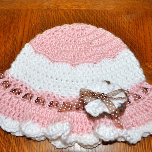 May include: A pink and white crocheted sun hat with a brown ribbon and bow. The hat has a scalloped edge and is made with a textured stitch.