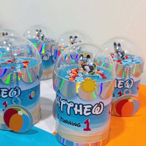 Puede incluir: Un recipiente cilíndrico para recuerdos de fiesta con tapa de cúpula transparente, con una envoltura temática de playa azul y el texto "MATTHEW IS TURNING 1". La parte superior contiene confeti colorido y un personaje, con un dije de pelota.