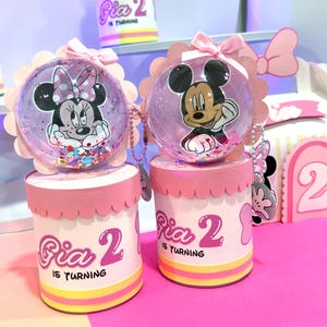 Puede incluir: Dos recipientes cilíndricos para recuerdos de fiesta con tapas festoneadas rosas y esferas de plástico transparente en la parte superior. Las esferas contienen confeti e imágenes de Minnie y Mickey Mouse. Los recipientes incluyen el texto "Gia 2 IS TURNING".