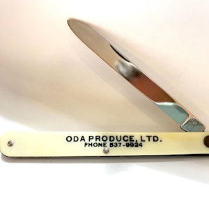 May include: A vintage folding knife with a shiny silver blade and a cream-colored handle. The handle has the text "ODA PRODUCE LTD. PHONE 537-9924" printed in black. The knife is partially open, displaying the blade.