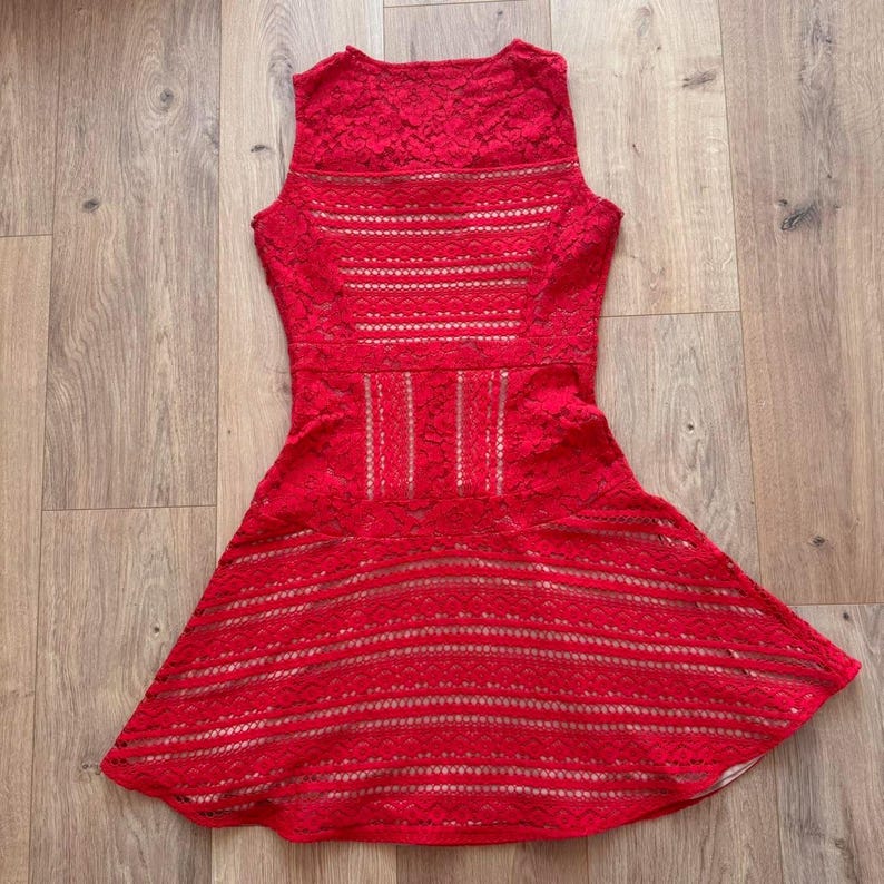 K&ouml;nnte beinhalten: Ein &auml;rmelloses rotes Kleid mit einer komplizierten Spitzenauflage und horizontalen Ausschnittmustern an Oberteil und Rock. Das Kleidungsst&uuml;ck hat eine taillierte Passform, die in einen ausgestellten A-Linien-Rock mit gewelltem Saum &uuml;bergeht.