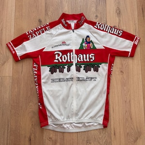 Puede incluir: Un maillot de ciclismo de manga corta en blanco y rojo con el logotipo de Rothaus, el texto Schwarzwald y un gráfico de una persona sosteniendo jarras de cerveza. La prenda incluye los textos Tannen Zäpfle y Biergit Kraft e ilustraciones.