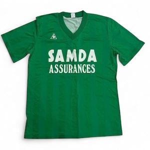 Puede incluir: Una camiseta verde de manga corta con cuello en V y rayas verticales tono sobre tono. El pecho muestra el texto blanco SAMDA ASSURANCES y un pequeño logotipo blanco de un gallo dentro de un triángulo en la parte superior izquierda.