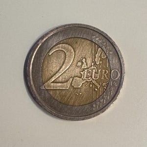 Puede incluir: Una moneda de dos euros con un anillo exterior plateado y un centro dorado. El número "2" se muestra a la izquierda, con la palabra "EURO" y un mapa de Europa a la derecha. Moneda de curso legal.
