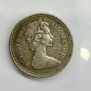 May include: A silver coin from 1983 depicting Queen Elizabeth II. The coin is round with a textured edge and features the inscription "ELIZABETH II" and the year. The coin's design includes a profile of the Queen.