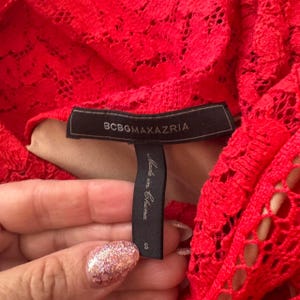 K&ouml;nnte beinhalten: Ein leuchtend rotes Kleidungsst&uuml;ck aus Spitze mit einem schwarzen Stoffetikett, das den Text BCBGMAXAZRIA in goldener Schrift zeigt. Ein darunter befestigtes schwarzes Etikett tr&auml;gt den Text Made in China und die Gr&ouml;&szlig;e S. Florales Muster auf beigem Futter.