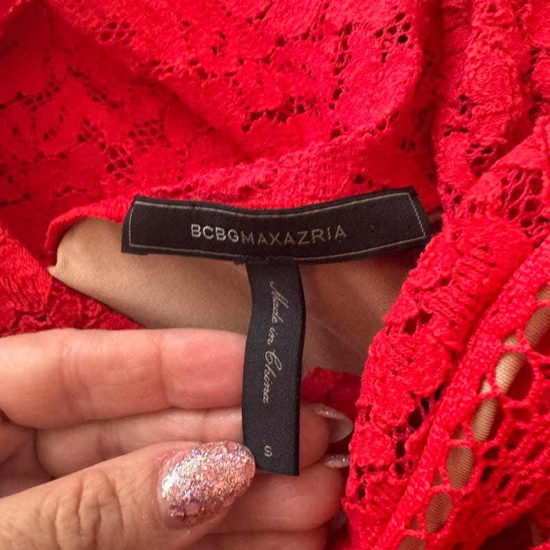 K&ouml;nnte beinhalten: Ein leuchtend rotes Kleidungsst&uuml;ck aus Spitze mit einem schwarzen Stoffetikett, das den Text BCBGMAXAZRIA in goldener Schrift zeigt. Ein darunter befestigtes schwarzes Etikett tr&auml;gt den Text Made in China und die Gr&ouml;&szlig;e S. Florales Muster auf beigem Futter.