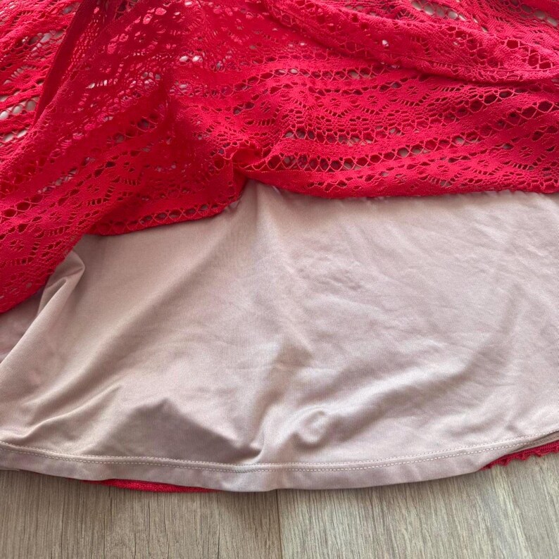 K&ouml;nnte beinhalten: Ein leuchtend rotes Kleidungsst&uuml;ck aus Spitze mit einem komplizierten, durchbrochenen Blumenmuster, das &uuml;ber einem soliden, hellbeigen, blickdichten Futter liegt. Der Spitzenstoff zeigt ein detailliertes, sich wiederholendes geometrisches und florales Design, w&auml;hrend die glatte beige Unterschicht einen Kontrast und Abdeckung unter dem leuchtend roten Textil bietet.