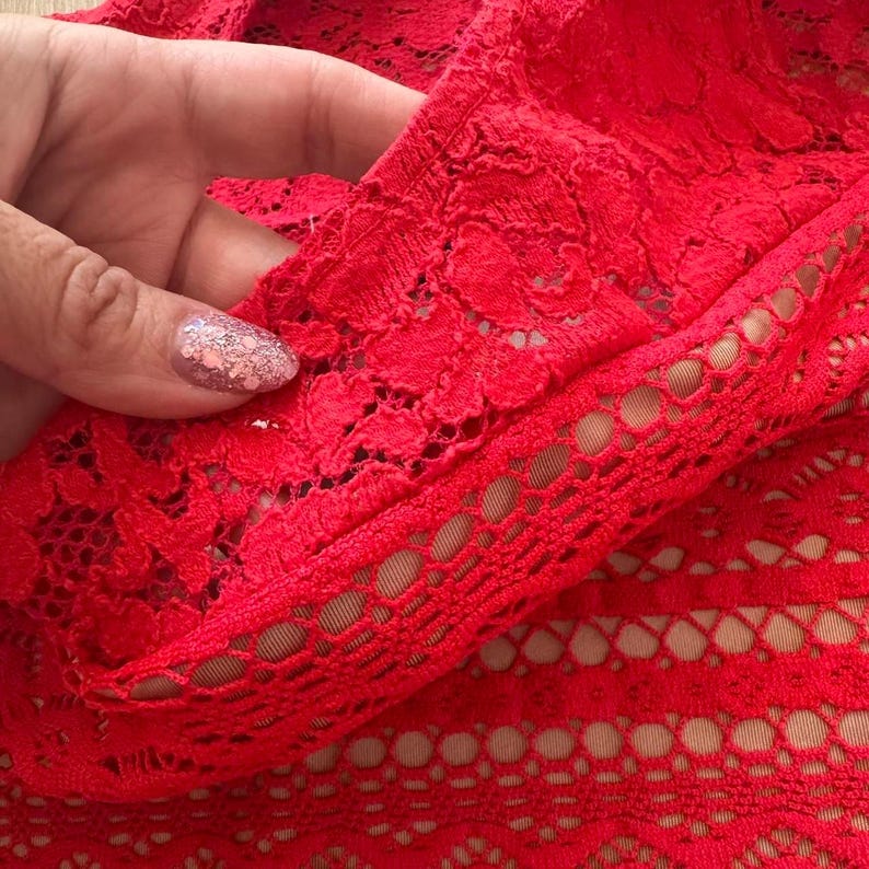 K&ouml;nnte beinhalten: Ein leuchtend roter Spitzenstoff mit einem detaillierten Blumenmuster und einem geometrischen Netzrand. Das Material ist halbtransparent und gibt den Blick auf ein darunter liegendes, neutralfarbenes Futter frei, das die Struktur betont.