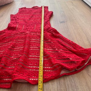K&ouml;nnte beinhalten: Ein &auml;rmelloses rotes Spitzenkleid mit beigem Futter, ausgestelltem Rock und Rundhalsausschnitt. Ein gelbes Ma&szlig;band liegt vertikal in der Mitte und zeigt eine Gesamtl&auml;nge von etwa 89 Zentimetern von der Schulter bis zum Saum des Kleides an.
