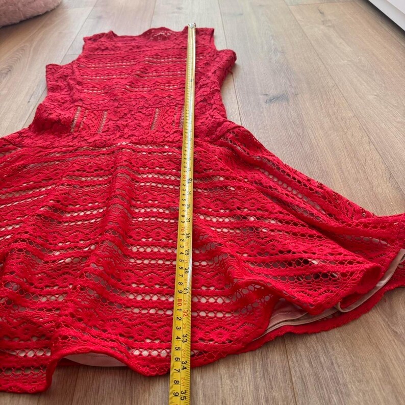 K&ouml;nnte beinhalten: Ein &auml;rmelloses rotes Spitzenkleid mit beigem Futter, ausgestelltem Rock und Rundhalsausschnitt. Ein gelbes Ma&szlig;band liegt vertikal in der Mitte und zeigt eine Gesamtl&auml;nge von etwa 89 Zentimetern von der Schulter bis zum Saum des Kleides an.