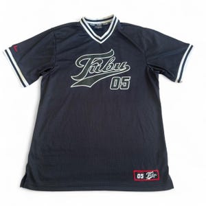 May include: A navy blue short-sleeve jersey with a V-neck collar featuring white stripes. The front displays the text Fubu and the number 05 in white with dark outlines. A small red and white patch with the text 05 Fubu is on the bottom hem.