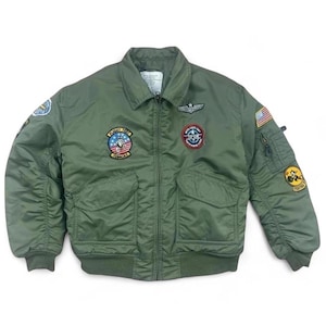 May include: Olive green bomber jacket with various patches, including a US flag and military insignia. It has a zip-up front, two front pockets, and ribbed cuffs and hem. The jacket has a classic military-inspired design.