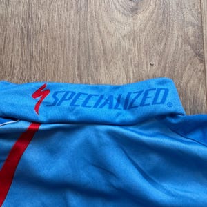 Może przedstawiać: Zbliżenie niebieskiego kołnierza koszulki sportowej z napisem "SPECIALIZED" w kolorze niebieskim i czerwonym logo błyskawicy. Koszulka ma czerwony pasek z boku.