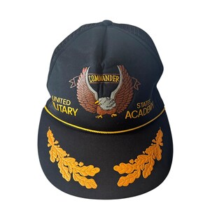 Può includere: Cappellino da baseball nero con retro in rete e visiera curva. Il cappellino presenta un disegno di aquila ricamata con la parola "COMMANDER" sopra. Le parole "UNITED MILITARY STATES ACADEMY" sono ricamate anche sul cappellino. Disegni di foglie dorate sulla visiera.