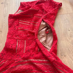 K&ouml;nnte beinhalten: Ein &auml;rmelloses rotes Spitzenkleid mit einem komplexen Blumenmuster und horizontalen B&auml;ndern aus verschiedenen Texturen. Das Kleidungsst&uuml;ck hat ein beiges Innenfutter, das an der offenen Seite sichtbar ist und einen roten Rei&szlig;verschluss zeigt.