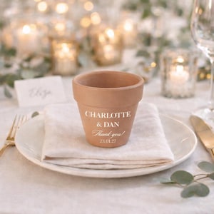 May include: A small, terracotta-colored flower pot with the names "CHARLOTTE & DAN" and the words "Thank you!" printed on the front. The pot sits on a white napkin and plate, part of a table setting.