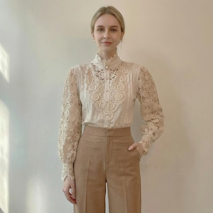 以下が含まれることがあります： 袖と胴体に繊細なレースのディテールが施されたクリーム色のブラウス。 ブラウスは、ハイネックのフリルカラーが特徴で、小さな真珠のようなボタンで前面がボタン留めされています。 ハイウエストのタンカラーのズボンと組み合わせています。