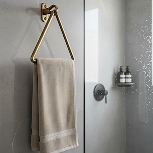 Op de afbeelding: Een driehoekige handdoekring van messing gemonteerd aan een muur, met een gevouwen beige handdoek met een getextureerd oppervlak en horizontale gestreepte details aan de onderkant. De metalen houder heeft een warme metallic afwerking.