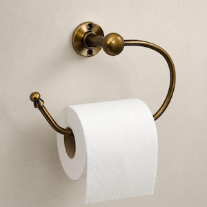 May include: A wall-mounted toilet roll holder with an antique brass finish. The holder features a circular mounting plate with two screw holes and a curved, C-shaped arm ending in a decorative ball finial, holding a roll of white toilet paper.