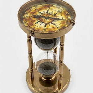 Op de afbeelding: Een messing zandloper in vintage-stijl met een kompas. Het kompas heeft richtingmarkeringen en een gedetailleerde wijzerplaat. De zandloper bevat donker zand en wordt ondersteund door een messing frame, een klassiek nautisch decor.