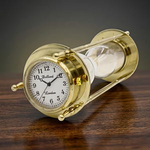 May include: A brass hourglass with a clock face, featuring the name "Dollond London." The clock face displays Roman numerals and a seconds dial. The hourglass is filled with white sand and is set on a wooden surface.