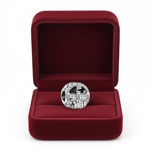 May include: A silver charm with a detailed depiction of a building, possibly a landmark, set inside a red velvet jewelry box. The charm has intricate architectural details and a flag. The box is open, revealing the charm.