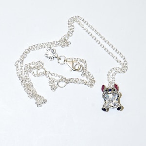 Op de afbeelding: Zilverkleurige ketting met een hanger van een stripfiguur met een helder kristallen lichaam, rode oren en blauwe accenten. De ketting heeft een fijne ketting en een bloemvormige sluiting.