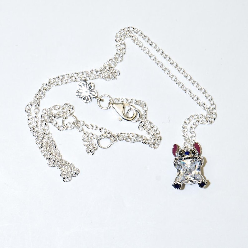 Op de afbeelding: Zilverkleurige ketting met een hanger van een stripfiguur met een helder kristallen lichaam, rode oren en blauwe accenten. De ketting heeft een fijne ketting en een bloemvormige sluiting.