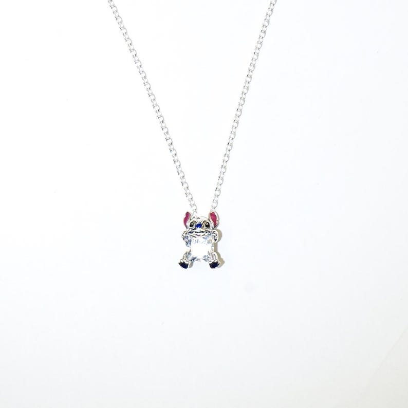 Op de afbeelding: Een zilveren ketting met een hanger van het personage Stitch. De hanger is een heldere, facetgeslepen kristal met blauwe, roze en zwarte details. De ketting is een fijne zilveren schakelketting.