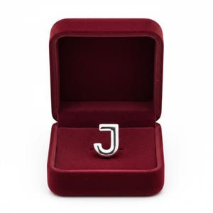 May include: A silver letter "J" ring inside an open, burgundy velvet ring box. The ring is a capital letter "J" with a rounded serif design. The ring box is square with a hinged lid and a soft, plush interior.