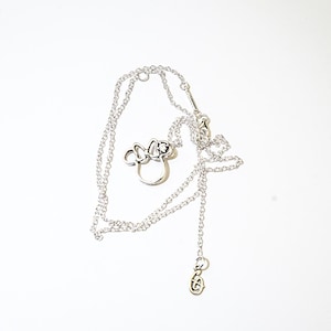 Puede incluir: Un collar plateado con un colgante con forma de cabeza y lazo de un personaje de dibujos animados. El collar tiene una cadena delicada y un peque&ntilde;o dije en el extremo. El colgante presenta una forma de cabeza circular con un lazo.